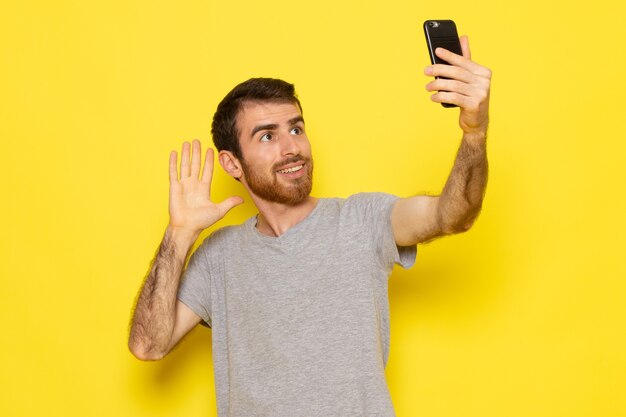 黄色の壁の男式感情カラーモデルでselfieを取って灰色のtシャツの正面の若い男性