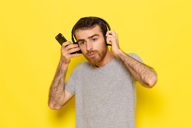 전화를 사용하고 노란색 벽 남자 컬러 모델에서 음악을 듣고 회색 티셔츠에 전면보기 젊은 남성