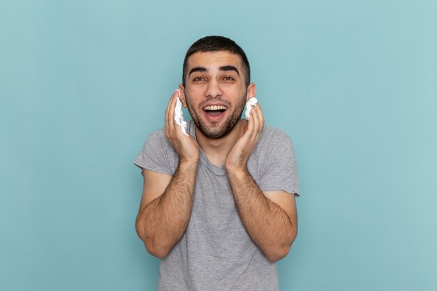 Front view young male in grey t-shirt using white foam for shaving on ice-blue