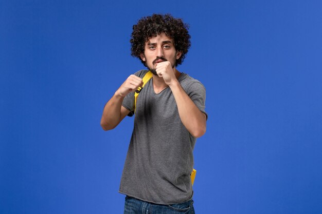 Front view of young male in grey t-shirt wearing yellow backpack on the blue wall