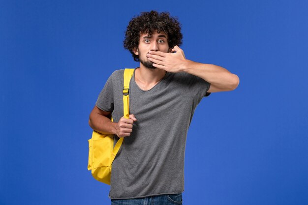 Front view of young male in grey t-shirt wearing yellow backpack closing his mouth on blue wall