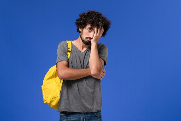 青い壁に落ち込んで黄色のバックパックを身に着けている灰色のTシャツの若い男性の正面図