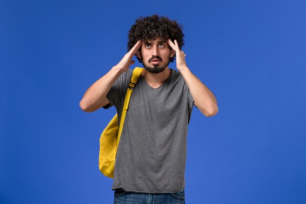 Вид спереди молодого мужчины в серой футболке в желтом рюкзаке, держащего виски на синей стене