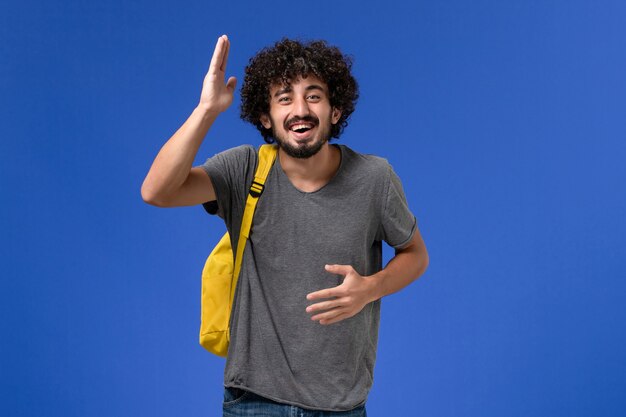 Front view of young male in grey t-shirt wearing yellow backpack laughing on the blue wall