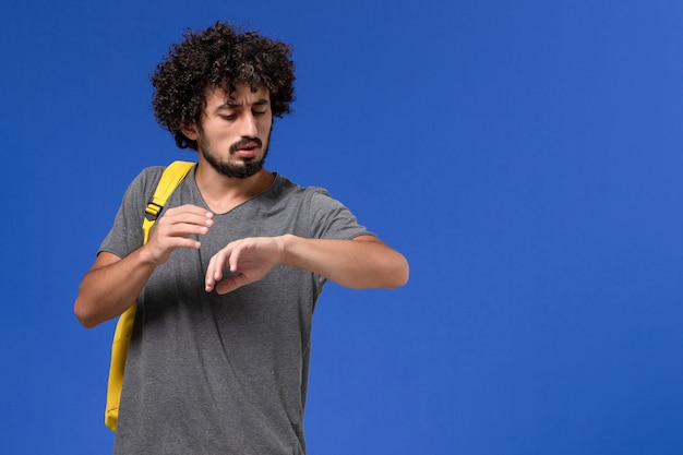 Free photo front view of young male in grey t-shirt wearing yellow backpack looking at his wrist on the blue wall