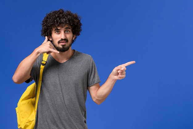 Front view of young male in grey t-shirt wearing yellow backpack posing on blue wall