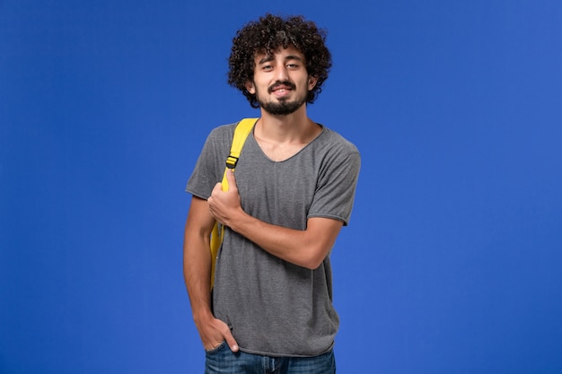 Вид спереди молодого мужчины в серой футболке с желтым рюкзаком, слегка улыбаясь на синей стене
