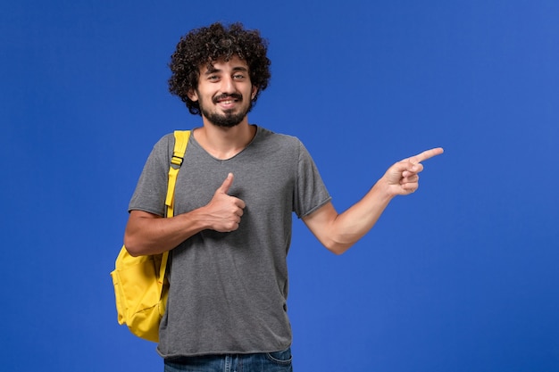 Vista frontale del giovane maschio in maglietta grigia che porta zaino giallo sorridente sulla parete blu