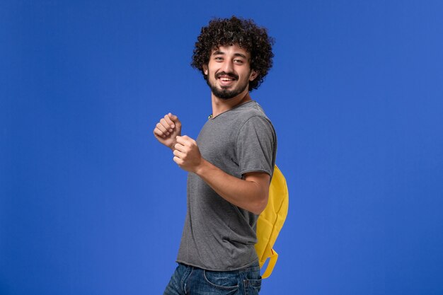 青い壁に笑みを浮かべて黄色のバックパックを身に着けている灰色のTシャツの若い男性の正面図