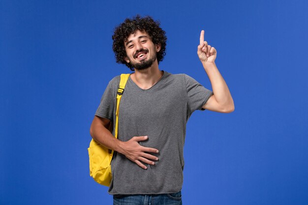 Вид спереди молодого мужчины в серой футболке с желтым рюкзаком, улыбающегося на синей стене