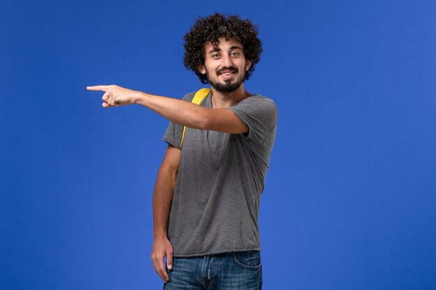 Front view of young male in grey t-shirt wearing yellow backpack smiling on the blue wall