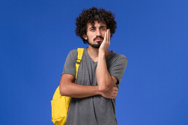 Front view of young male in grey t-shirt wearing yellow backpack thinking on the blue wall