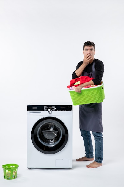 Front view of young male holding basket with dirty clothes on white wall