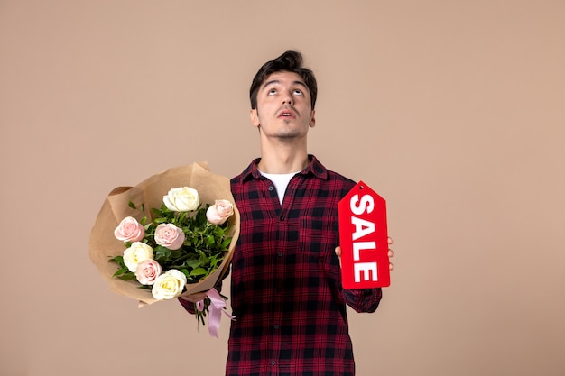 Free photo front view young male holding beautiful flowers and sale nameplate on brown wall