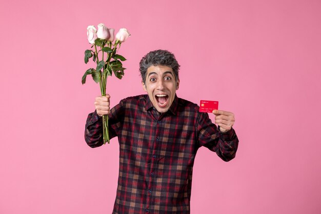 Front view young male holding beautiful pink roses and bank card on pink wall