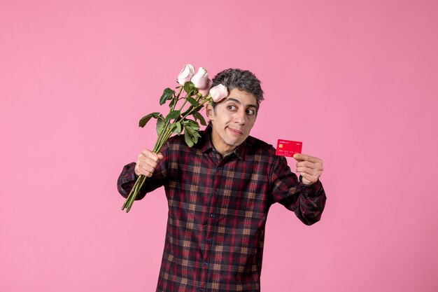 Front view young male holding beautiful pink roses and bank card on pink wall