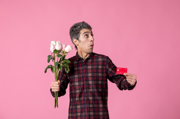 Вид спереди молодой мужчина держит красивые розовые розы и банковскую карту на розовой стене