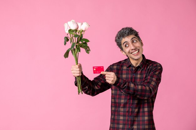 Front view young male holding beautiful pink roses and bank card on pink wall