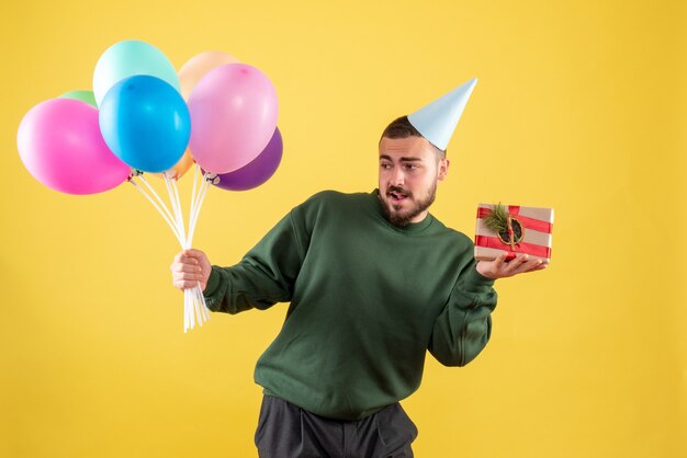 Front view young male holding colorful balloons and present on yellow background