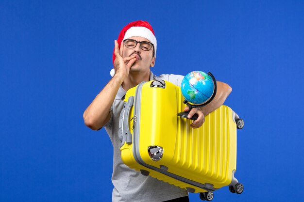 Вид спереди молодого мужчины, держащего глобус и желтую сумку на синей стене, самолеты, отпуск