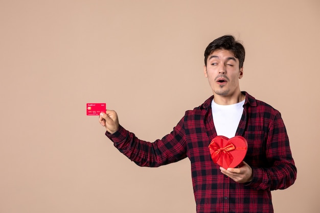 Вид спереди молодой мужчина держит подарок в форме сердца и банковскую карту на коричневой стене