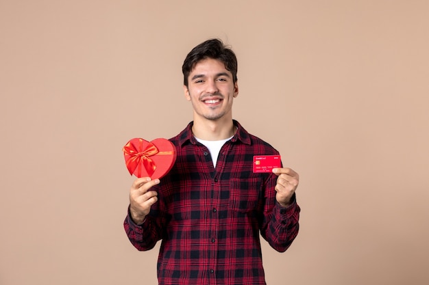 Front view young male holding heart shaped present and bank card on brown wall