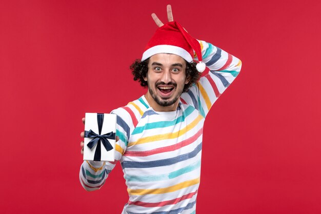 Вид спереди молодой мужчина держит праздничный подарок на красной стене новогодний праздник красные эмоции