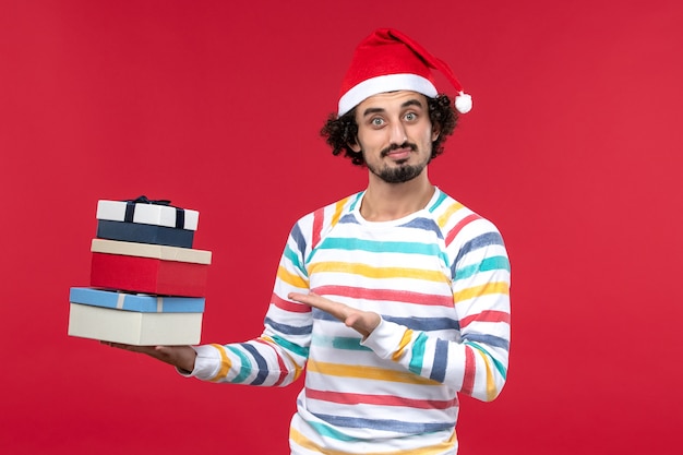 Вид спереди молодой мужчина держит праздничные подарки на красной стене новогодний праздник эмоции