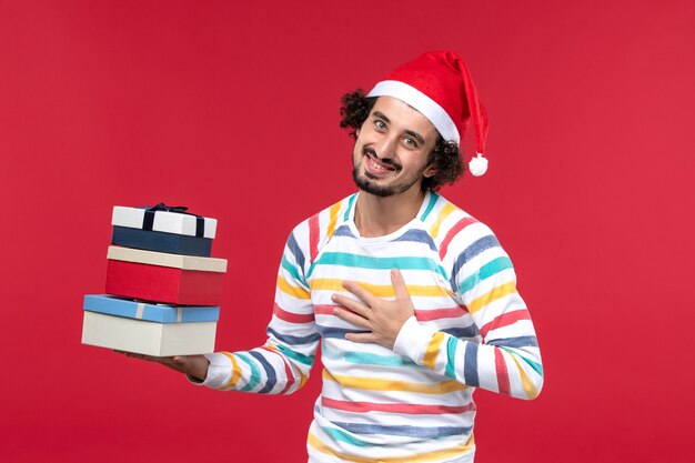 Вид спереди молодой мужчина держит праздничные подарки на красной стене новогодние праздничные эмоции