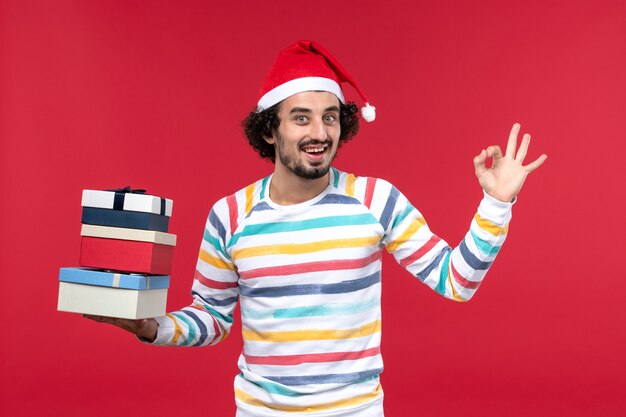 Front view young male holding holiday presents on a red wall new year holidays emotion