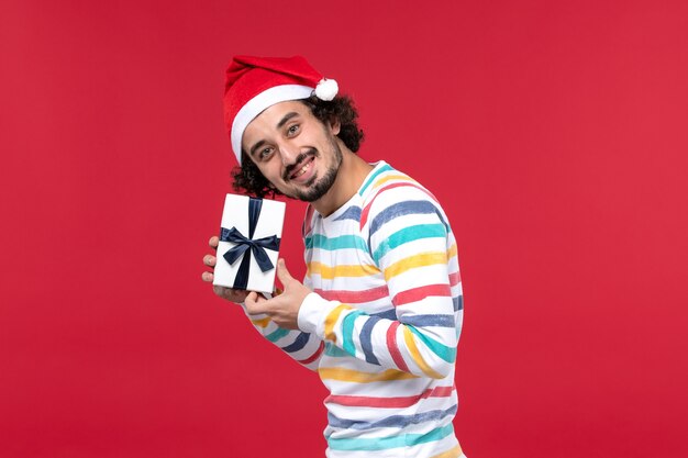 Вид спереди молодой мужчина держит новогодний подарок на красном фоне праздник новогодняя эмоция