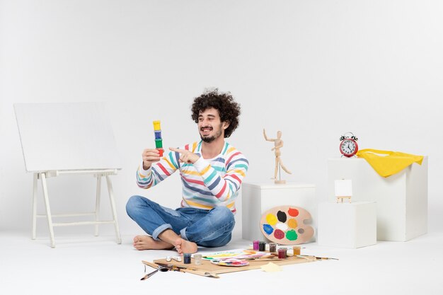 Front view of young male holding paints for drawing inside little cans on white wall