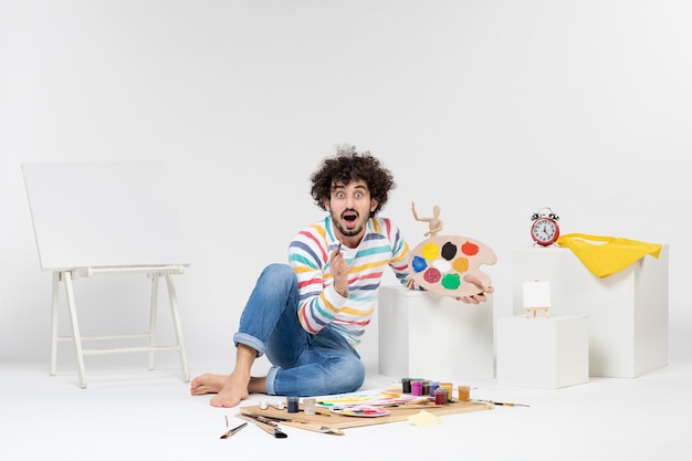 Вид спереди молодого мужчины, держащего краски и кисточку для рисования на белой стене