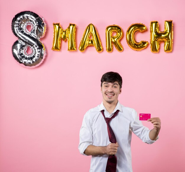 분홍색 배경에 3월 장식이 있는 분홍색 은행 카드를 들고 있는 전면 보기 젊은 남성 파티 선물 휴가 남자 여성 돈 평등 색상 쇼핑