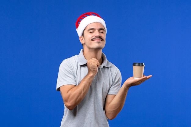 Вид спереди молодого мужчины, держащего пластиковую кофейную чашку на синем столе, эмоция мужского нового года