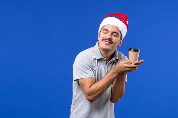 Вид спереди молодого мужчины, держащего пластиковую чашку кофе на синей стене, эмоции мужского нового года