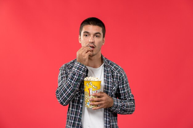 Front view young male holding popcorn package eating it on light-red wall cinema theater film male movie