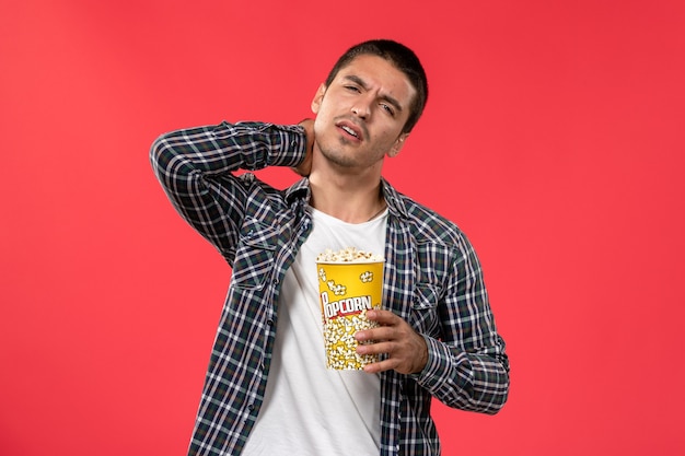 Front view young male holding popcorn package and having neckache on red surface cinema theater film movie