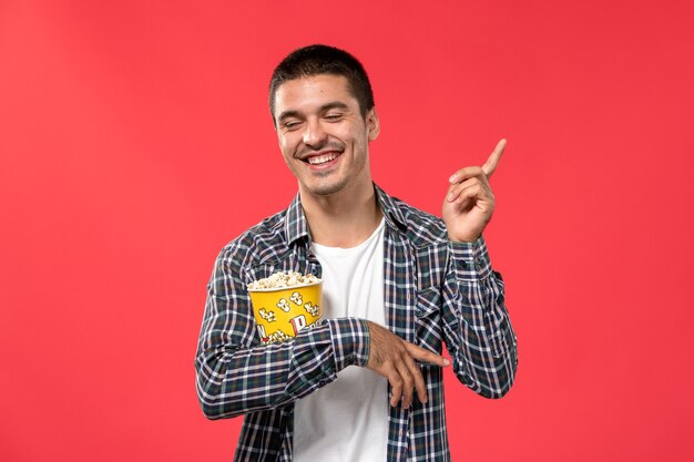 Front view young male holding popcorn package and laughing on red surface cinema theater film movie