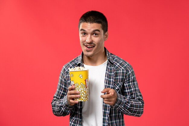 Front view young male holding popcorn package and posing on the red surface cinema theater film movie