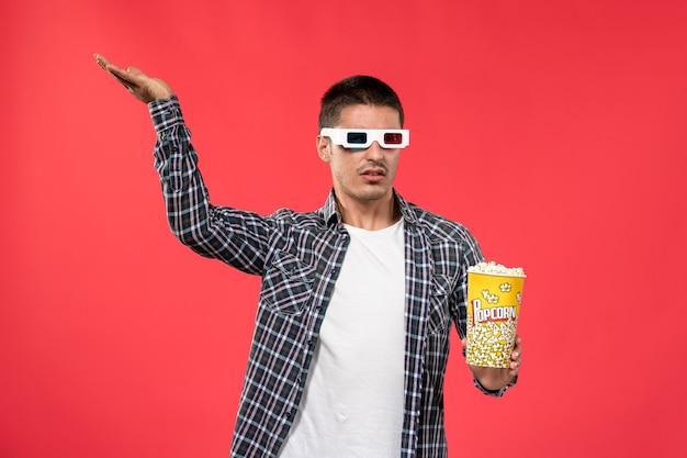 Front view young male holding popcorn package and wearing -d sunglasses on light-red wall cinema movie theater film