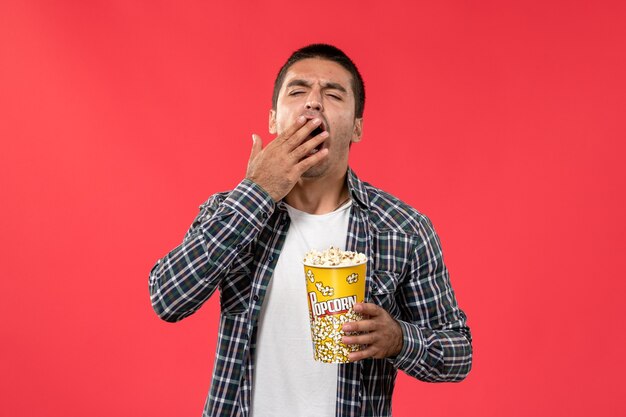 Front view young male holding popcorn package and yawning on red wall cinema theater film movie