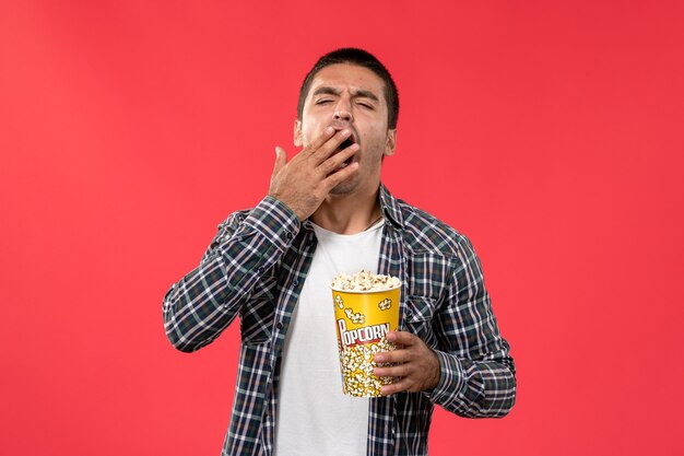 ポップコーンパッケージを保持し、赤い壁の映画館の映画館の映画映画であくびをしている正面図若い男性