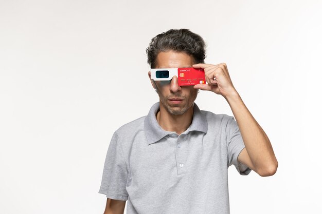 Front view young male holding red bank card in d sunglasses on a white surface