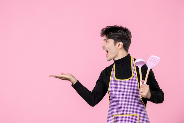 Вид спереди молодой мужчина в плаще держит розовые ложки на розовом фоне профессия еда работа приготовление еды тесто униформа цвет горизонтальный