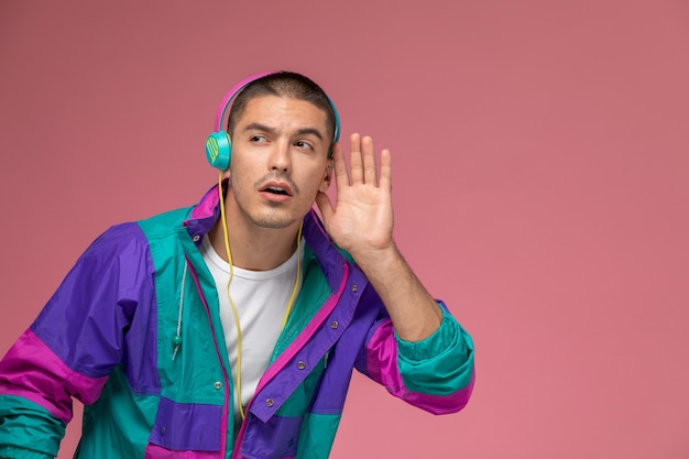 Бесплатное фото Вид спереди молодой самец в красочном пальто слушает музыку, пытаясь услышать на розовом фоне
