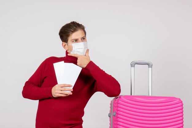 Front view of young male in mask holding plane tickets on a white wall