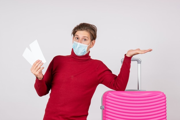 Front view of young male in mask holding plane tickets on the white wall