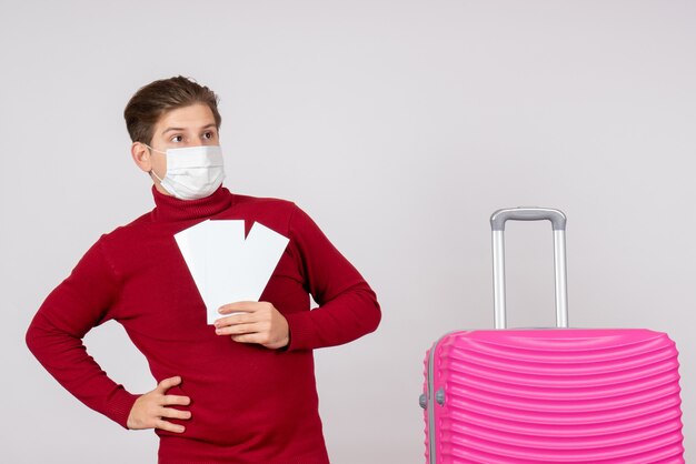 Front view of young male in mask holding plane tickets on white wall