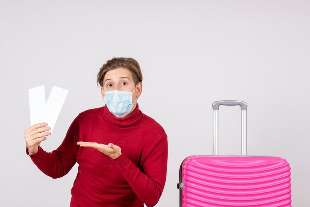 Front view of young male in mask holding plane tickets on white wall